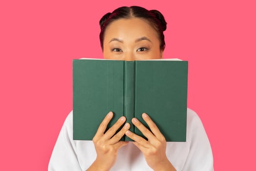 Asian woman joyfully holds a green book in front of her face, with a playful expression, set against a vibrant pink background. photo