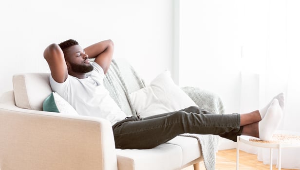 Black man sits on a white couch with his arms behind his head, relaxing and enjoying a moment of peace. His feet are up on a small table beside the couch, and he appears to be taking a break photo