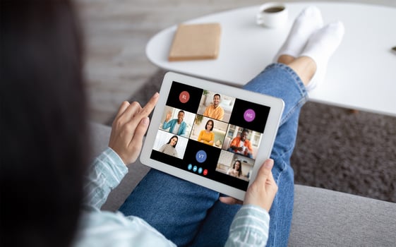 A woman sits on a couch, her feet resting on a coffee table, while holding a tablet. The screen displays a conference with multiple participants. photo