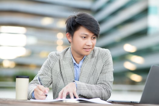 A young Asian man sits at a table outside an office building, working on his laptop. He is writing in a notebook with a pen, and his laptop is open in front of him. photo