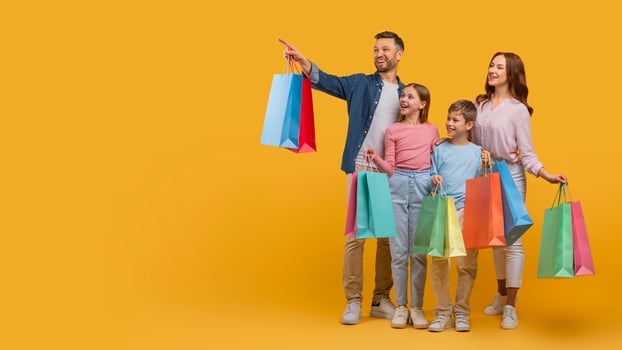 A joyful family, with two parents and two children, are standing together holding colorful shopping bags. The father points out something in the distance, copy space photo