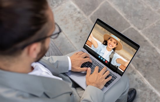 A man sits outdoors on a tiled surface, using his laptop to chat with a woman. He is wearing a gray suit jacket and glasses, and she is wearing a hat, a blue shirt, and a backpack photo