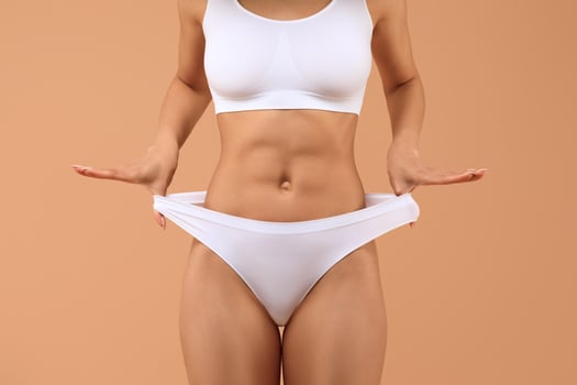 Cropped of woman wearing white underwear and a white sports bra poses with her hands on her hips against a tan background. Her arms are extended to the sides, cropped photo