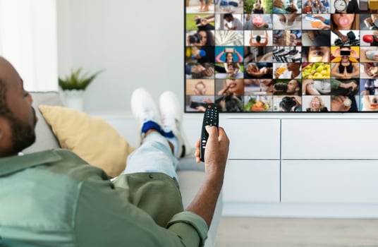 Black man is relaxing on a couch in his living room, watching television with a remote control in his hand. He is wearing casual clothing and has his feet up on the couch. photo