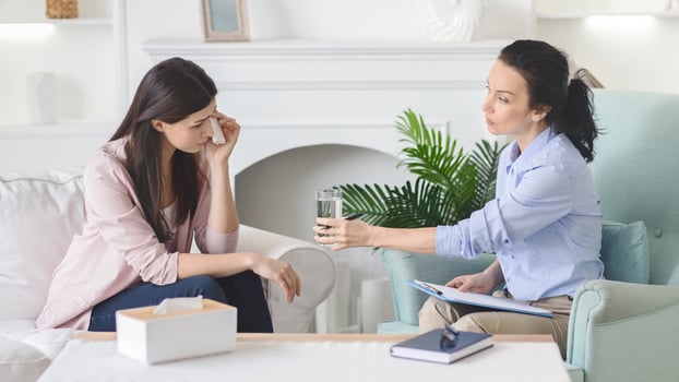 A therapist extends a glass of water to a distressed woman in a cozy living room setting during a daytime counseling session. The woman, showing visible signs of distress photo