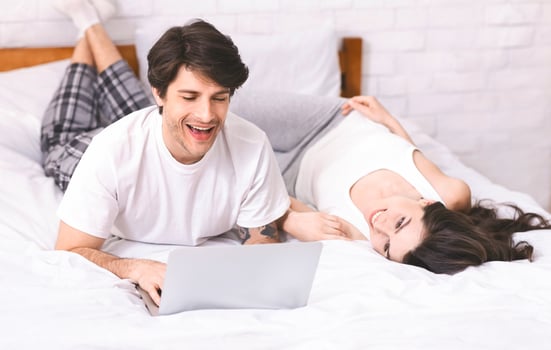 A man and a woman are relaxing in bed together. The man is using a laptop and smiling while looking at the screen. The woman is lying on her back with her head turned to look at him, smiling. photo