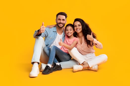 A happy family of three, a mother, father, and young daughter, sit on a yellow background, smiling and giving thumbs up. The family is dressed in casual clothes, they are all looking at the camera. photo