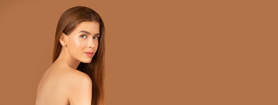 Feminine nude woman with silky skin and long straight hair posing on brown studio background. Charming young lady pampering herself, looking at camera, taking care of her body, copy space photo