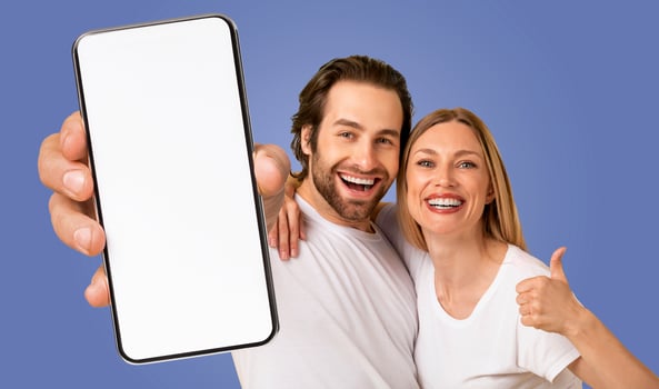 A happy couple stands in front of a blue background, holding a smartphone with a blank screen in front of them. The man is smiling and pointing at the phone, while the woman is giving a thumbs up photo
