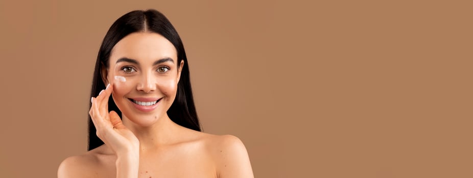 Cheerful sensual naked long-haired brunette millennial woman with smile on her face using facial eye cream for young smooth glowing skin, isolated on beige studio background, copy space photo