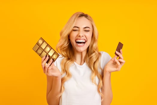 A young woman with long blonde hair is standing in front of a bright yellow background. She is wearing a white t-shirt and holding two chocolate bars, one in each hand. photo