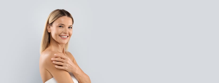Spa treatment concept. Cheerful smiling middle aged half-naked blonde woman embracing her shoulders, looking at camera, copy space for advertisement, isolated on grey studio background photo
