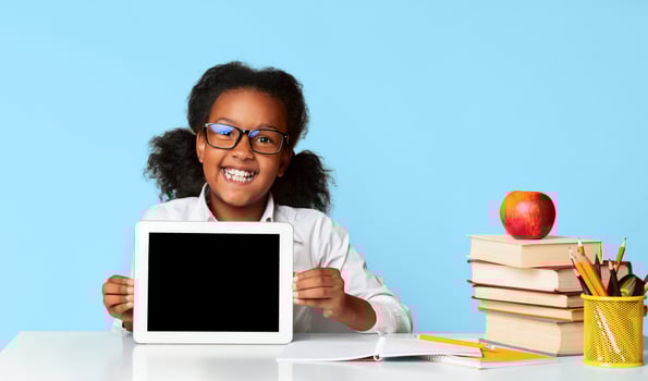 African American Schoolgirl Showing Tablet Blank Screen Sitting At Desk On Yellow Studio Background. School And Educational Apps. Mockup photo