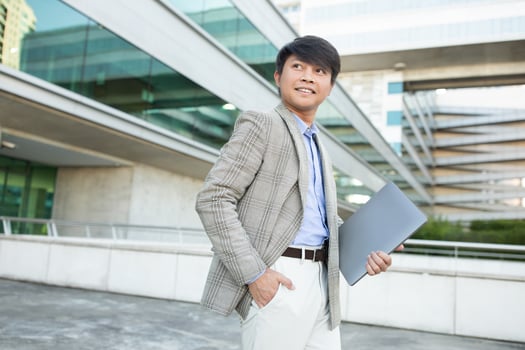 Asian man in a blazer and white pants walks outside a modern office building, holding a laptop and looking towards the right. The building has large windows and a sleek, contemporary design. photo