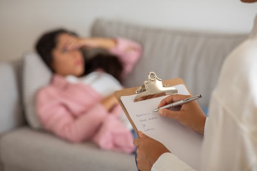 A therapist sits with a clipboard and pen, taking notes while a patient lies on a couch in the background. The patient appears to be distressed and is out of focus. photo