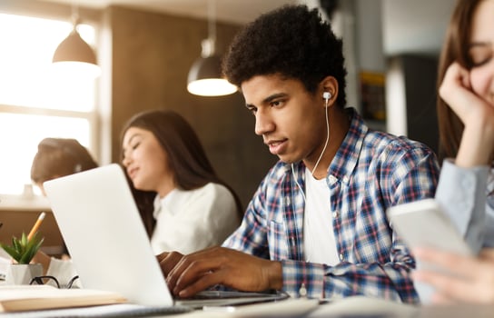 African American young man is engrossed in his work on a laptop in a bustling office setting. He wears earphones, likely immersed in music or an audio call, as he types intently photo
