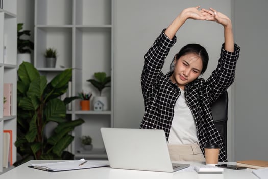 Young Female Accountant Stretching at Desk, Tired from Work in Modern Office Setting with Laptop and Documents photo