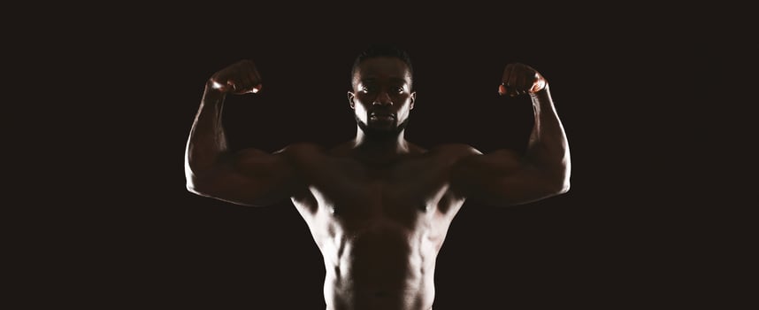 A muscular man flexes his biceps in a dark studio setting, with only his body illuminated. photo