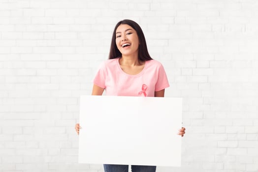Breast Cancer Cure. Happy Asian Girl In Pink T-Shirt With Ribbon Holding Empty Poster Standing On White Background. Mockup photo