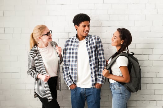 A diverse group of friends are standing in front of a white brick wall, looking towards the camera. They are casually dressed, chatting and laughing, with varied expressions photo