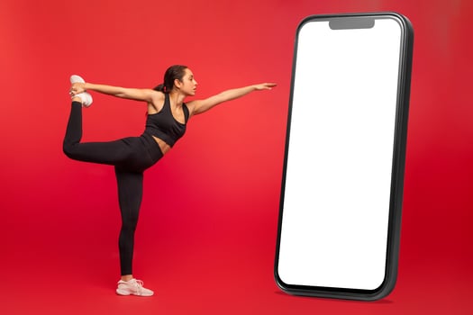 Flexible young woman in black sportswear exercising next to huge phone with white blank screen, on red studio background, practicing yoga or pilates, full length photo