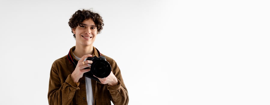 A young man with curly hair stands in a bright studio, holding a camera and smiling. The backdrop is light-colored, providing a professional atmosphere for photography, copy space photo