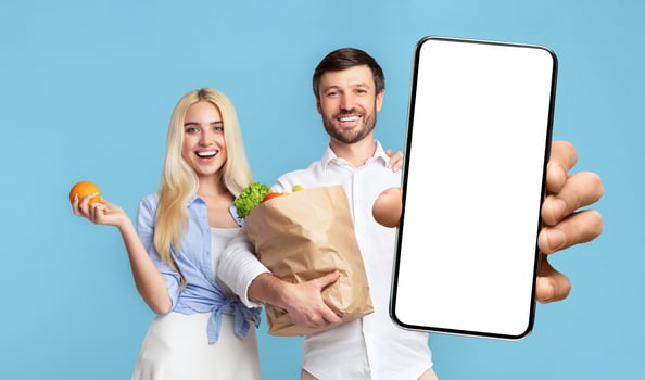 A man and woman stand side by side, both smiling at the camera. The woman is holding an orange and the man is holding a paper bag full of groceries and a smartphone with a blank white screen photo
