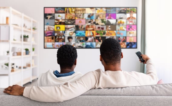 Two men are relaxing on a couch in a living room, watching television. The television is displaying a large, colorful grid of images. photo
