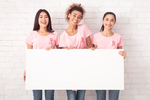 Three Multiracial Breast Cancer Volunteers Girls Holding Blank White Board Posing Against Brick Wall. Mockup photo