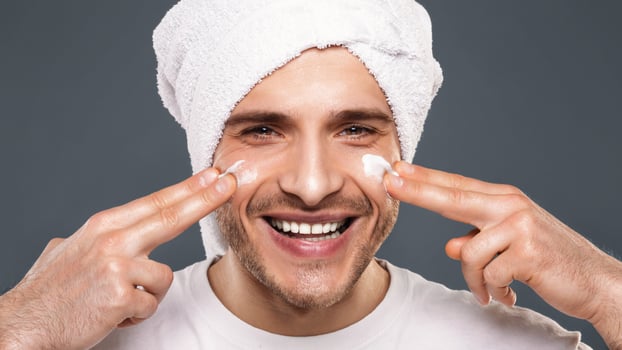 Morning hygiene. Millennial guy with towel on his head, applying cream on face, smiling at camera photo