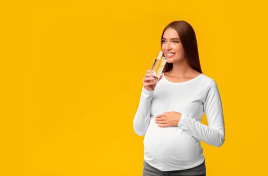 Cheerful Pregnant Girl Drinking Water From Glass Standing In Studio Over Pink Background. Free Space For Text photo