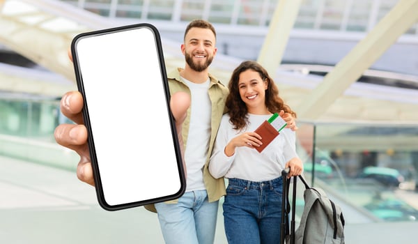 A beaming young man and woman stand inside a bustling airport terminal, showcasing a large, blank smartphone screen. photo