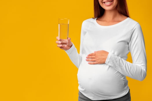 Unrecognizable Pregnant Lady Holding Glass Of Water Touching Belly Standing On Pink Background In Studio. Cropped, Copy Space photo