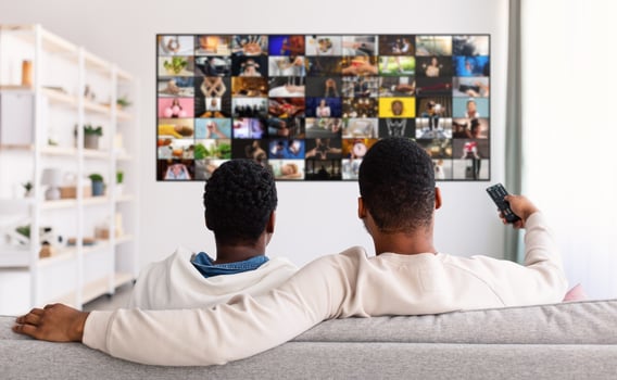 Two men sit on a gray couch watching television in a living room. The man on the left is wearing a white hoodie and the man on the right is holding a remote control photo