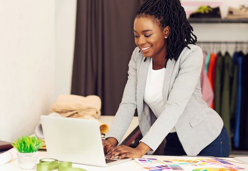 A smiling African American woman fashion designer strikes a pose with a measuring tape in her workshop, garments all around, using laptop photo