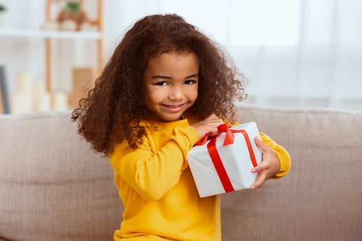 Kid's Birthday. Cute African Little Girl Unpacking Wrapped Gift Sitting With Mother On Sofa At Home. photo