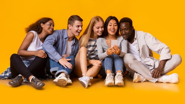 Group of multiracial cheerful classmates having break, watching videos on smartphone over yellow background, panorama photo