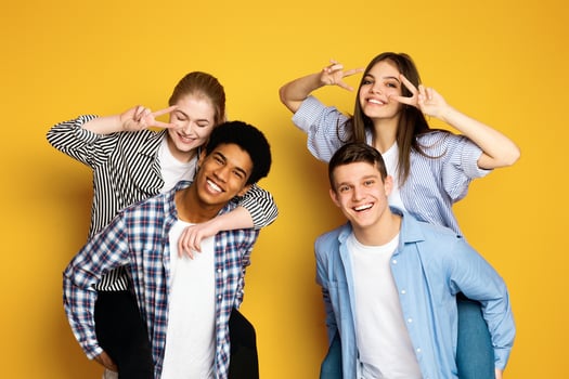 A cheerful group of four young friends is playfully posing together, with two making peace signs near their eyes and all sporting genuine, joyful smiles. photo