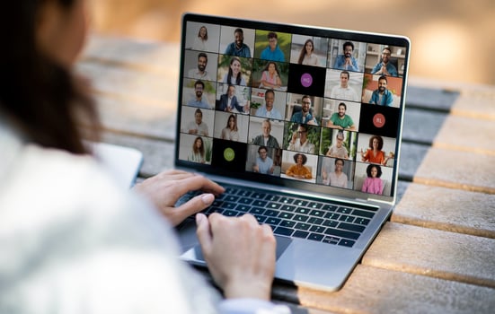Lady sitting at an outdoor table, actively engaging in a virtual business meeting on a laptop. The screen displays numerous participants in different windows. photo
