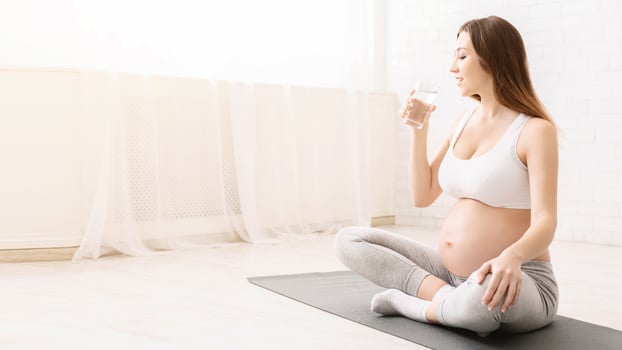 A pregnant woman is shown sitting on a yoga mat and drinking water. She looks relaxed and engaged in her hydration routine. photo