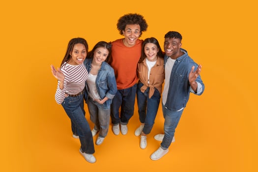 A group of five friends, all dressed in casual attire, pose together on a bright yellow background. They look happy and excited, with their arms around each other, view above photo