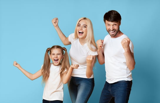 A family of three, a father, mother, and daughter, stand together in front of a blue background, all three raising their fists in the air and smiling excitedly. photo