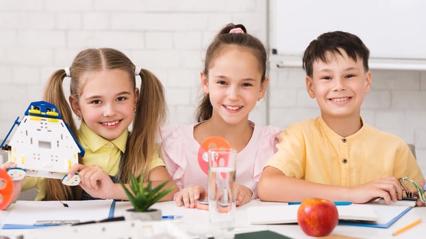 Three children, a girl holding a robot and two others, are sitting at a table in a classroom setting, smiling and working on a STEM project. photo
