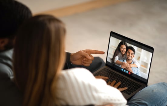 A couple is sitting on a couch at home, chatting with their friends on a laptop. The man is pointing at the screen, while the woman is smiling and looking at the laptop photo