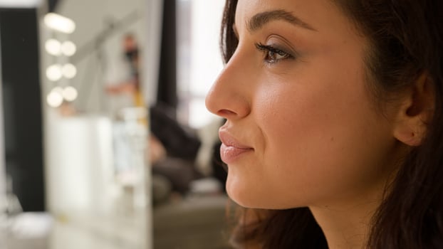 Glad cheerful Caucasian woman model girl applying nude lipstick lips doing make-up beauty routine. Female lady use decorative cosmetic product smile looking at mirror profile view indoors blog studio photo
