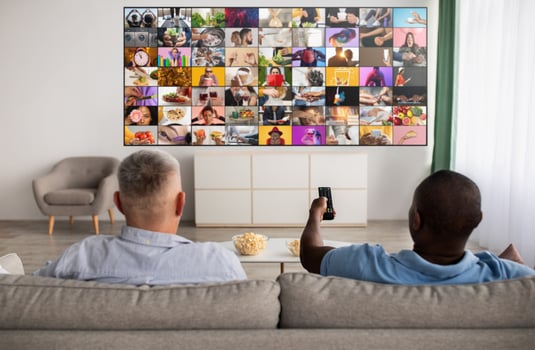 Two men are relaxing on a couch in a living room. They are watching television. The television screen is displaying a collage of many different images. One man is holding a remote control. photo