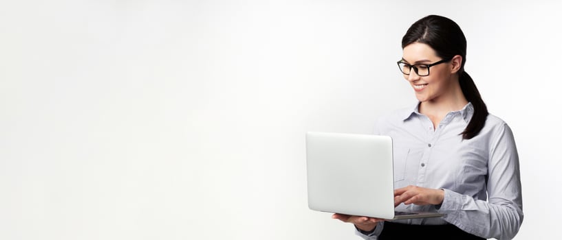 A woman wearing glasses smiles as she uses a laptop in a white studio setting. photo