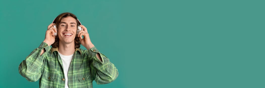 Music everywhere. Cheerful young guy with headset listening to favorite song on turquoise studio background, panorama with copy space photo