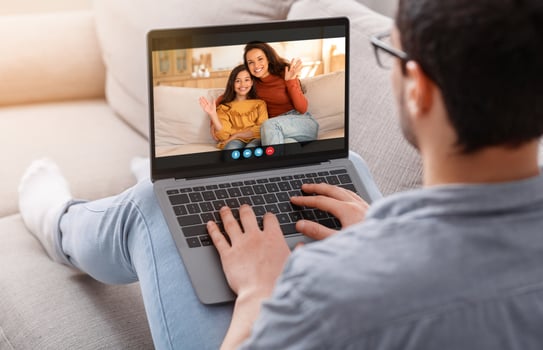 A man sits on a couch, wearing casual clothing and glasses, using a laptop to chat with his wife and daughter. The woman and girl are sitting together, waving at the man. photo