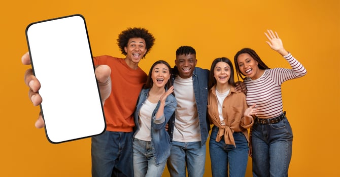 Five multiethnic friends stand together, smiling and posing enthusiastically, with one holding up a blank smartphone. The cheerful atmosphere is highlighted by a vibrant yellow backdrop. photo